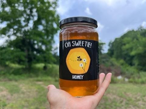 A person holding a jar of honey labeled "Oh Sweetie!" with a vibrant design featuring a bee and flowers, set against a natural outdoor background. The honey is golden and clear, showcasing its quality.