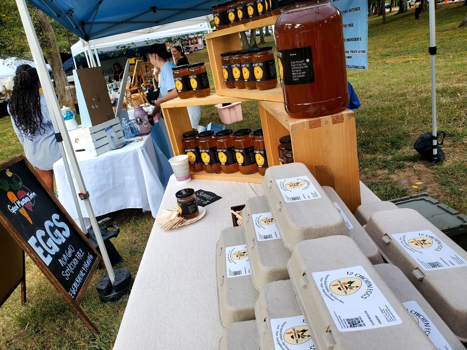 Farmers market stall featuring fresh chicken eggs in eco-friendly packaging, jars of honey, and a sign promoting egg sales. The setup includes wooden shelves and a variety of local produce, highlighting organic and sustainable farming practices.